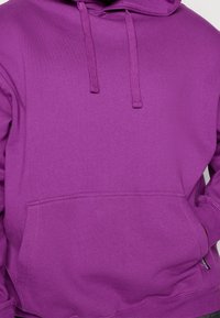 Violetti huppari, jossa kengurutasku, hupun nyörit ja resorireunukset hihoissa. Sileä, lämmin kangas ja yksivärinen malli.