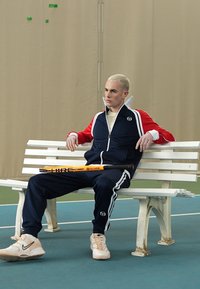 Tamsiai mėlynas sportinis kostiumas su raudonais akcentais ir baltais dryžiais, sėdintis ant suolo, laiko oranžinį tenisą raketę. Jaukus audinys, sportiškas dizainas.