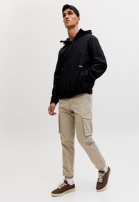 Junger Mann trägt eine schwarze Kapuzenjacke mit Reißverschluss, beige Cargo-Hosen, weiße Socken und braune Turnschuhe, steht vor einem einfachen weißen Hintergrund.