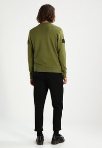 Olivově zelená mikina s dlouhými rukávy, žebrovanými manžetami a lemem; na obou ramenou má černé látkové nášivky. Nošena s černými kalhotami.