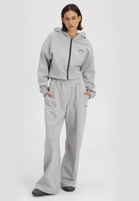 Grijze cropped hoodie met een frontale rits en zwart logo; gecombineerd met hooggewaiste, loszittende grijze sweatpants met zakken. Zacht materiaal.