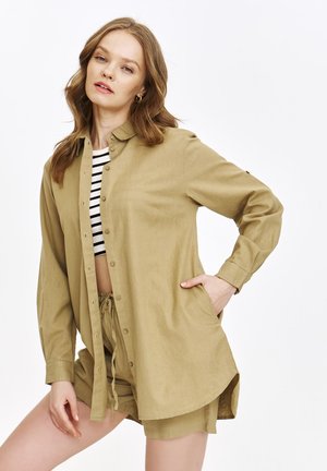 Khaki koszula zapinana na guziki o luźnym kroju, z długimi rękawami i dołem, który opada po bokach, noszona na czarno-białą, pasekową crop top.