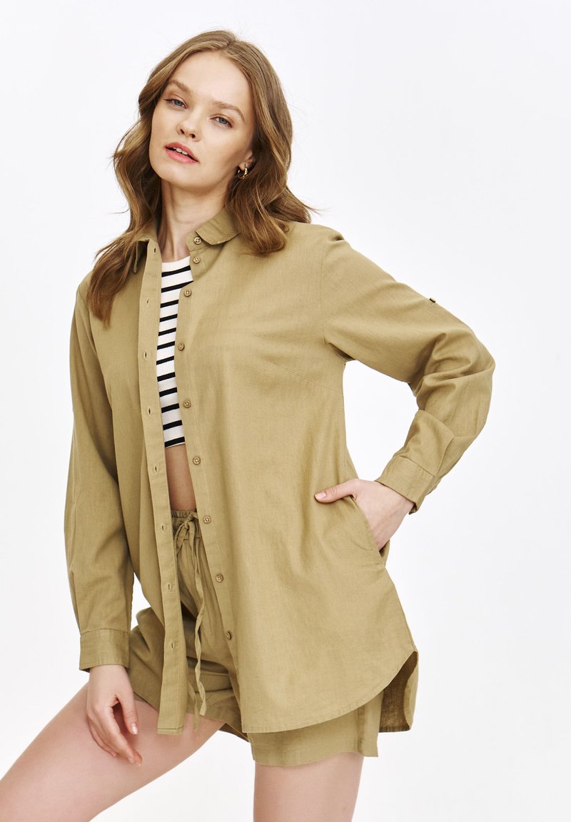 Khaki koszula zapinana na guziki o luźnym kroju, z długimi rękawami i dołem, który opada po bokach, noszona na czarno-białą, pasekową crop top.