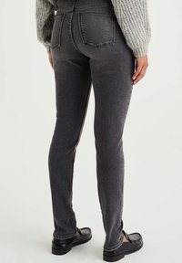 Hochtaillierte, dunkelgraue Skinny-Jeans mit einer glatten Textur, ausgestattet mit zwei Gesäßtaschen und subtilen Nähten. Kombiniert mit schwarzen Slippern.