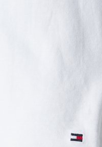 T-shirt en coton blanc avec un petit logo de drapeau brodé en rouge, blanc et bleu dans le coin inférieur gauche. Texture douce avec un fin côtelè.