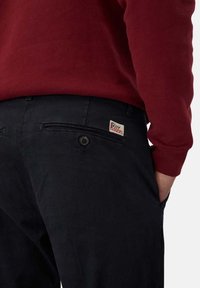 Pantaloni neri con una superficie liscia, due tasche anteriori e una tasca posteriore con un'etichetta cucita. Abbinati a una camicia a maniche lunghe color maroon.