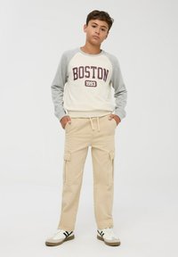Felpa crema con maniche raglan grigie che presenta una grafica "BOSTON 1993", abbinata a pantaloni cargo beige con tasche laterali e sneakers.