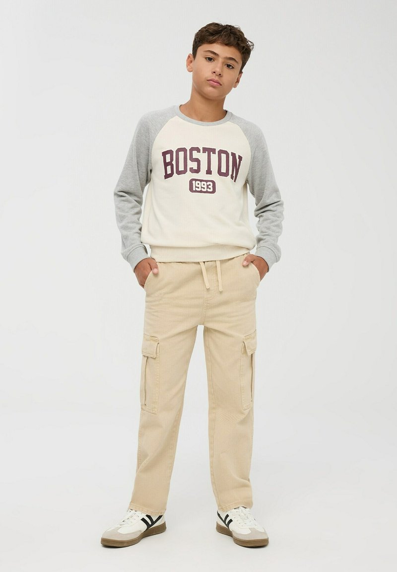 Felpa crema con maniche raglan grigie che presenta una grafica "BOSTON 1993", abbinata a pantaloni cargo beige con tasche laterali e sneakers.