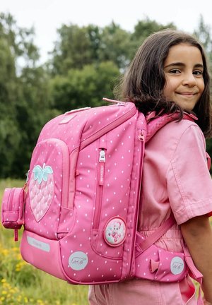 Ragazza sorridente all'aperto che indossa una maglietta rosa e porta uno zaino grande rosa decorato con fragole e piccoli puntini bianchi.