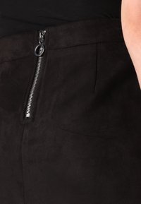 Svarta mockabyxor med dragkedja fram och en silverfärgad metallslider samt en slät, texturerad yta.