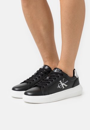 Black leather sneakers with a white rubber sole, featuring black laces and a silver logo. The back has a silver accent detail.