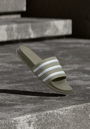 Olivengrøn Adidas slide sandal med tre hvide striber, der svæver over betontrappen i udendørs omgivelser.