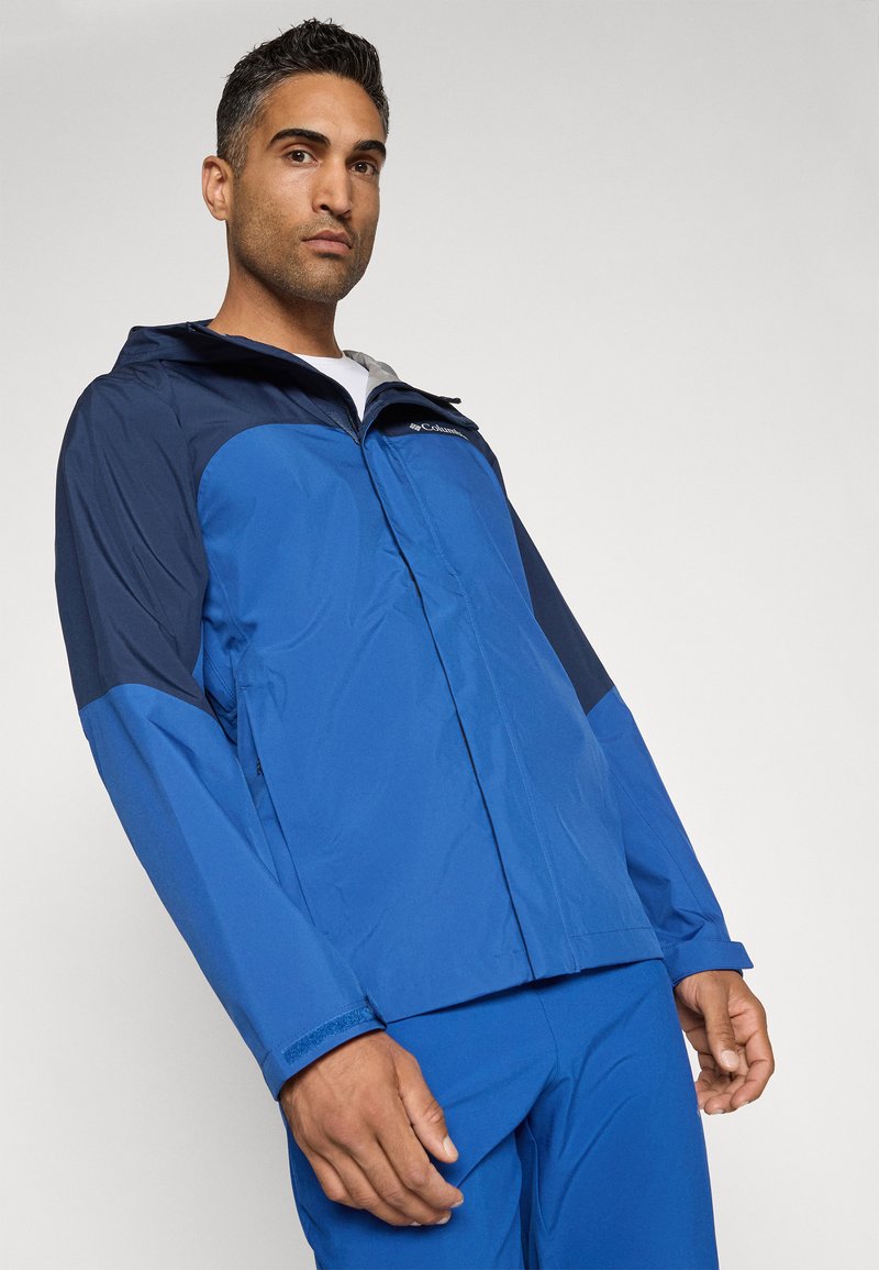 Niebieska wodoodporna kurtka z granatowymi akcentami, z zamkiem błyskawicznym, regulowanym kapturem i elastycznymi mankietami. Lekka, gładka tekstura.