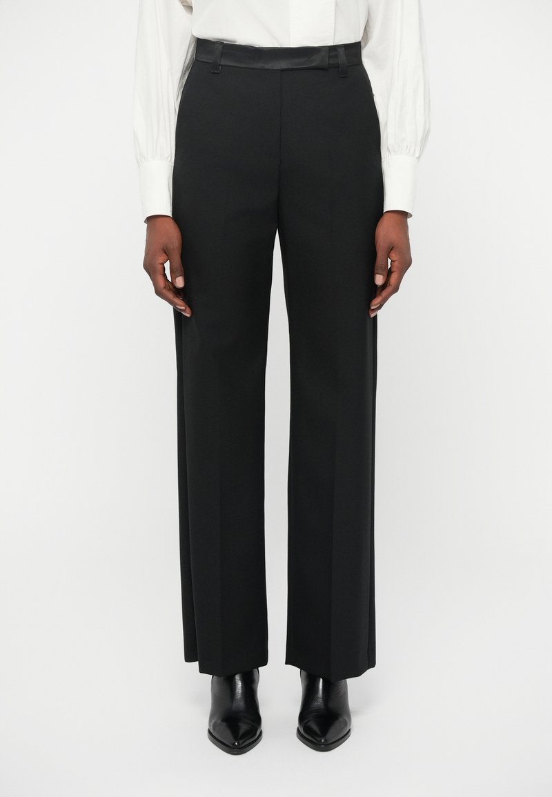 Musthave musta püksid sirge lõikega, millel on sile vöökoht ja tagasihoidlikud ees olevad voldid, kombineeritud terava musta kingaga.