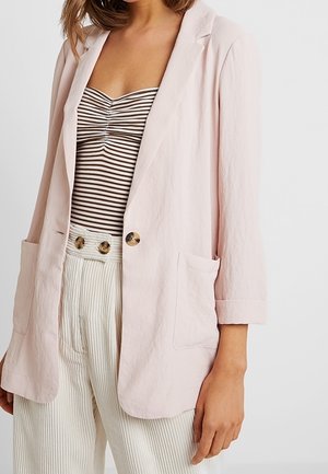 Femme portant un blazer rose clair sur un débardeur froncé à rayures marron et blanc, accompagné d'un pantalon blanc taille haute avec des détails à boutons.