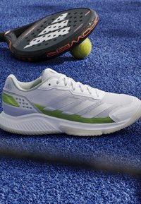 Scarpa sportiva bianca e verde su una superficie blu strutturata accanto a una racchetta da paddle nera e una palla da tennis gialla.