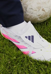 White soccer cleat with pink accents, featuring a sleek design, textured surface, and a distinctive logo. Grass and soccer ball in background.