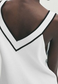 Weißes Tanktop mit V-förmigem Rücken, akzentuiert durch einen dünnen schwarzen Rand entlang des Halsausschnitts und der Träger, aus einem glatten Stoff gefertigt.