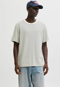 Person iført en navy cap, stribet hvid og sort t-shirt, og løse lyseblå jeans mod en ensfarvet baggrund.