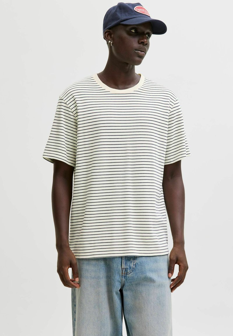 Person iført en navy cap, stribet hvid og sort t-shirt, og løse lyseblå jeans mod en ensfarvet baggrund.