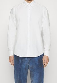 Man i en vit knappad långärmad skjorta och blå kordflorsbyxor, stående mot en enkel bakgrund.