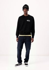 Sort sweatshirt med hvid grafik, lagd oven på en gul skjorte. Denim jeans og sorte sneakers med hvide accenter. Sort beanie på hovedet.