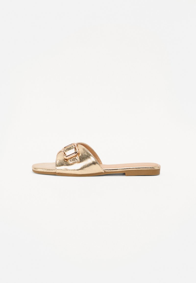 Sandalha metálica dourada com sola plana, apresentando um detalhe de fivela proeminente na parte superior. Textura suave com um design minimalista.