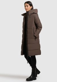 Braune Puffjacke mit Kapuze, ausgestattet mit einem Reißverschluss, horizontalem Steppmuster und Taschen an den Seiten. Trägt sie über schwarzen Leggings und Stiefeln.