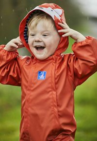 Imperméable orange avec un motif de renard sur la capuche, des poignets élastiques et une doublure rayée bleue, porté par un enfant en plein air.