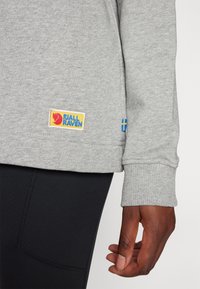 Grijze sweatshirt met geribde manchetten, voorzien van een Fjällräven logo patch in rood en blauw. Zwarte legging zichtbaar onder de sweatshirt.