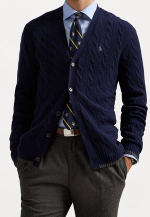 Man draagt een marineblauwe kabelgebreide cardigan over een blauw overhemd met een gestreepte crest-stropdas en grijze broek, handen in de zakken.