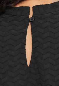 Black textured fabric top featuring a zigzag pattern, a keyhole cutout at the back, and a black button closure at the neckline.