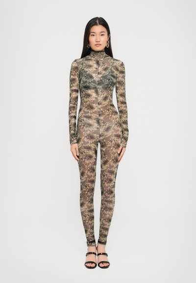 Kvinde står iført en gennemsigtig, tætsiddende blomstret bodystocking med lange ærmer, høj hals og sorte strop-sandaler mod en hvid baggrund.