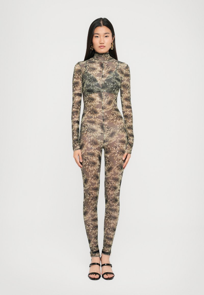 Mujer de pie con un body floral ajustado y transparente, de mangas largas, cuello alto y tacones negros con tiras, contra un fondo blanco.