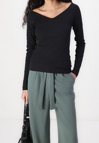 Schwarz geripptes Langarmoberteil mit V-Ausschnitt, kombiniert mit grünen plissierten Hosen. Das Oberteil hat eine figurbetonte Silhouette und eine weiche Textur.