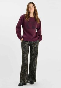 Burgundy sweatshirt with graphic print, paired with dark green leopard-print flared pants. Model stands with hands in pockets, on a plain background.