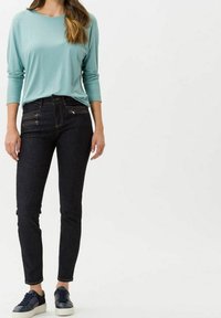 Blusa de manga larga azul clara combinada con jeans ajustados de mezclilla oscura con bolsillos con cremallera, llevada con zapatillas azules y blancas.