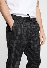 En person som bär en vit t-shirt och svarta rutiga byxor, som avslappnat har handen i fickan, står framför en neutral bakgrund.