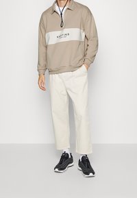 Beige zip-up sweatshirt med vit rand och svart text, ihop med ljusa vida byxor och svarta träningsskor.