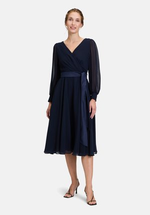 Marineblaues Wickelkleid mit langen, durchsichtigen Ärmeln, einem taillierten Oberteil und einem fließenden Rock. Mit einem passenden Gürtel, der in der Taille gebunden ist.