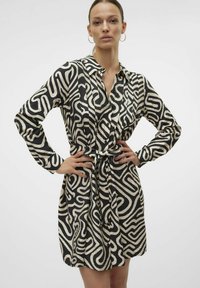 Abito camicia nero e beige con un motivo vorticoso, colletto, maniche lunghe e dettaglio di vita legata. Tessuto testurizzato.