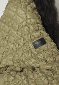 Gros plan sur une veste matelassée kaki avec un écusson noir à logo DKNY sur la manche, portée par une personne aux longs cheveux ondulés et foncés.