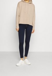 Beige Kapuzenpullover mit Kordelzügen, lockere Passform, kombiniert mit dunkelblauen Skinny Jeans und weißen Sneakers mit strukturierten Sohlen.
