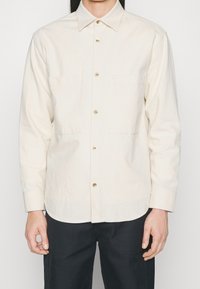 Chemise couleur crème à boutons avec col pointu, deux poches avant et boutons dorés. Fabriquée en tissu texturé.