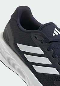 Gros plan sur une chaussure de sport noire avec une semelle blanche et trois bandes blanches sur le côté, avec des lacets noirs et un col rembourré.