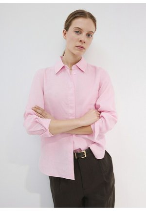Femme aux cheveux châtain clair portant une chemise rose pâle boutonnée et un pantalon noir avec ceinture, debout les bras croisés contre un mur blanc.
