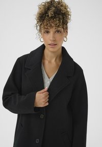 Woman with curly hair wearing gold hoop earrings and a black coat over a gray top, holding the coat lapel with one hand.