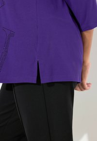 Violet lyhythihainen paita, jossa on koristeellinen kivisuunnittelu, sivuhalkio helmassa ja sileä, pehmeä tekstuuri. Yhdistetty mustiin housuihin.