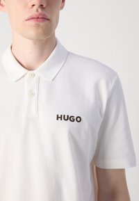 Hvit polo skjorte laget av bomull med en myk tekstur. Har en klassisk krage, to-knapps placket, og ordet "HUGO" i mørkebrun brodering.