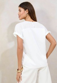 Weißes Blusenshirt mit aufgerollten Ärmeln und Knopfakzenten, mit einem Schlüssellochverschluss im Rücken und glatter Stoffoberfläche.